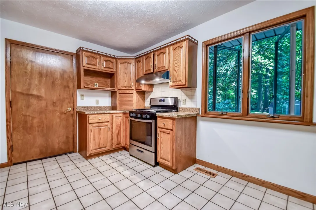 Kitchen with stainless steel gas stove, under cabinet range hood, backsplash, open shelves, and a textured ceiling