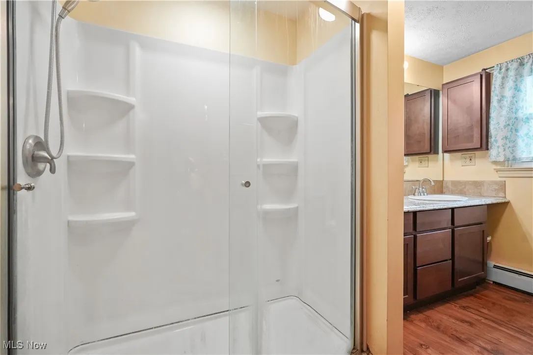 Full bath with a shower stall, wood finished floors, vanity, a textured ceiling, and a baseboard radiator