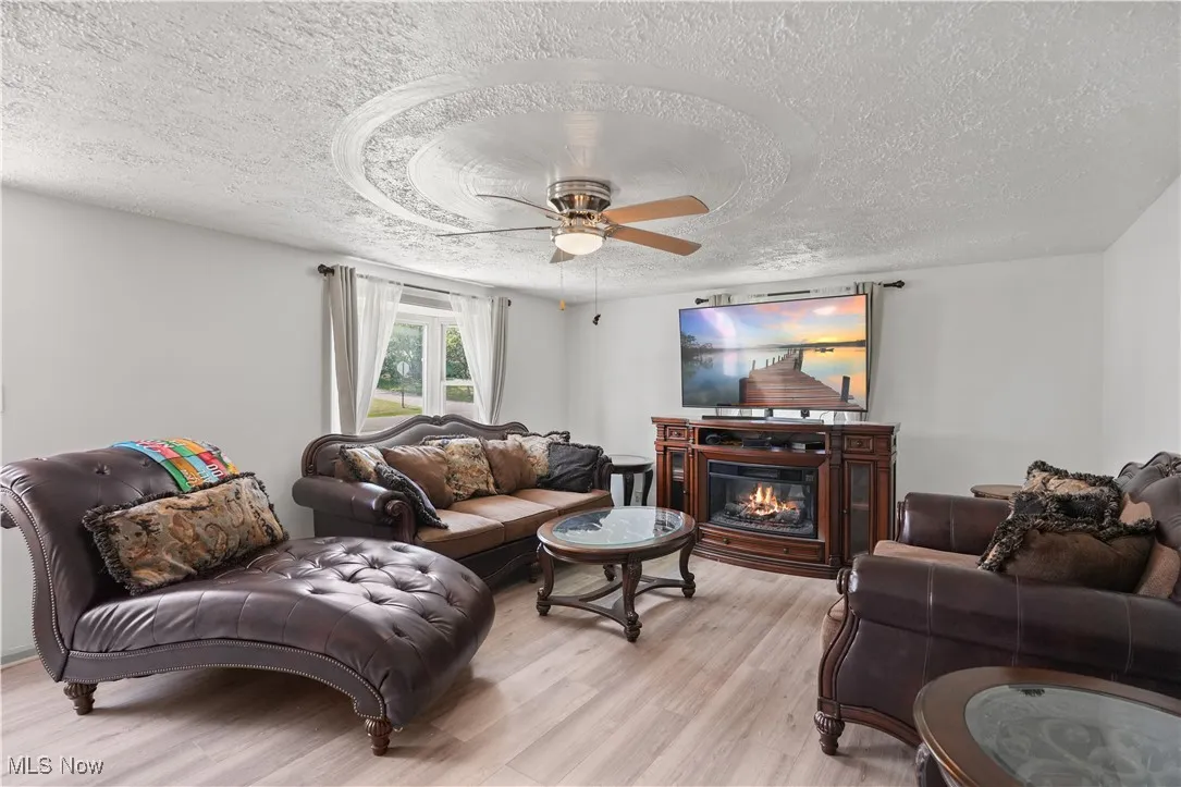 Living area featuring a textured ceiling, light wood-style flooring, a ceiling fan, and a glass covered fireplace