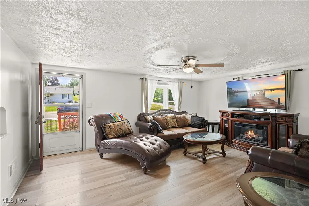 Living area with a textured ceiling, light wood-type flooring, a ceiling fan, and a glass covered fireplace