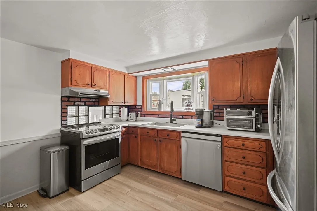 Kitchen featuring stainless steel appliances, light countertops, light wood finished floors, brown cabinetry, and under cabinet range hood