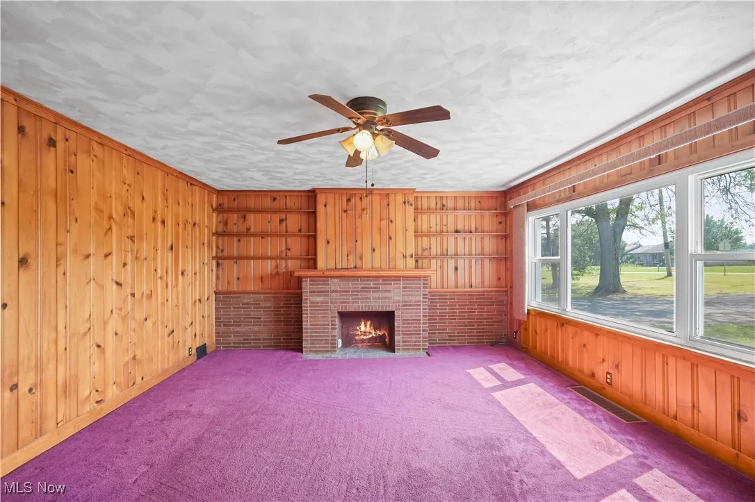 Unfurnished living room featuring wooden walls, a ceiling fan, carpet flooring, and a brick fireplace