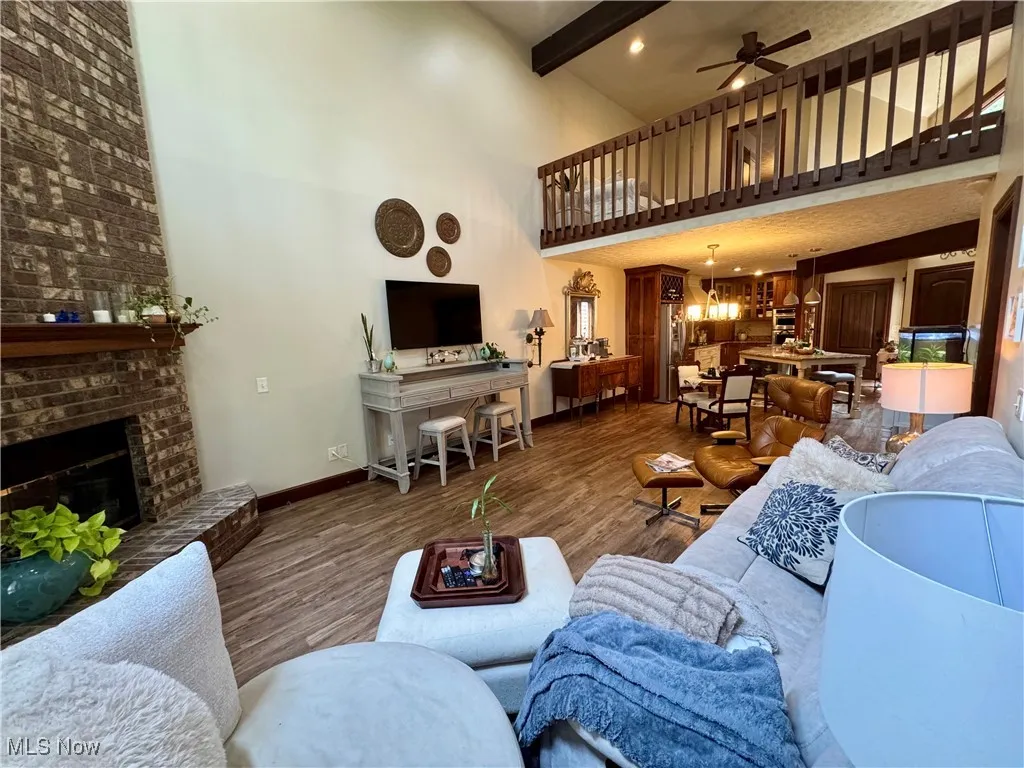 Living area with a towering ceiling, wood finished floors, beam ceiling, a ceiling fan, and a brick fireplace