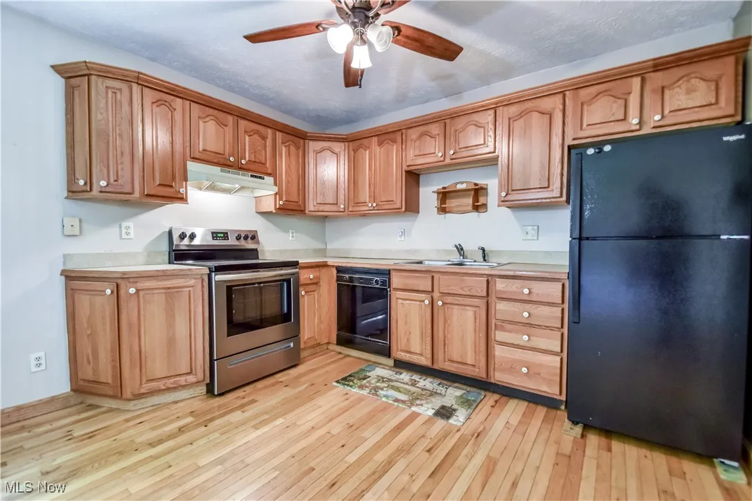 Kitchen featuring black appliances, light wood finished floors, under cabinet range hood, ceiling fan, and light countertops