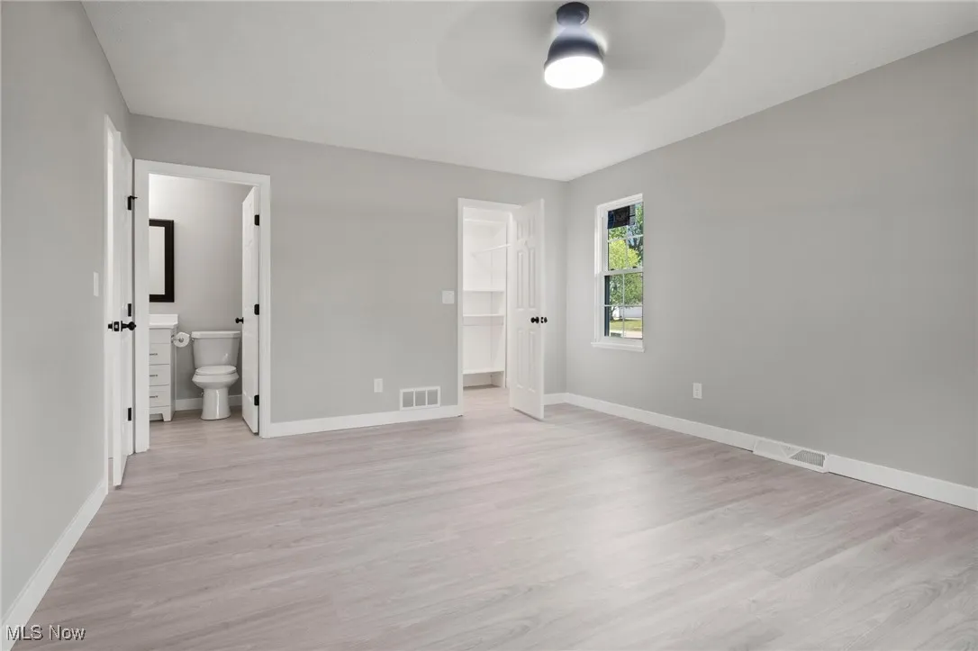 Unfurnished bedroom with light wood-style floors, a spacious closet, ensuite bathroom, and a ceiling fan