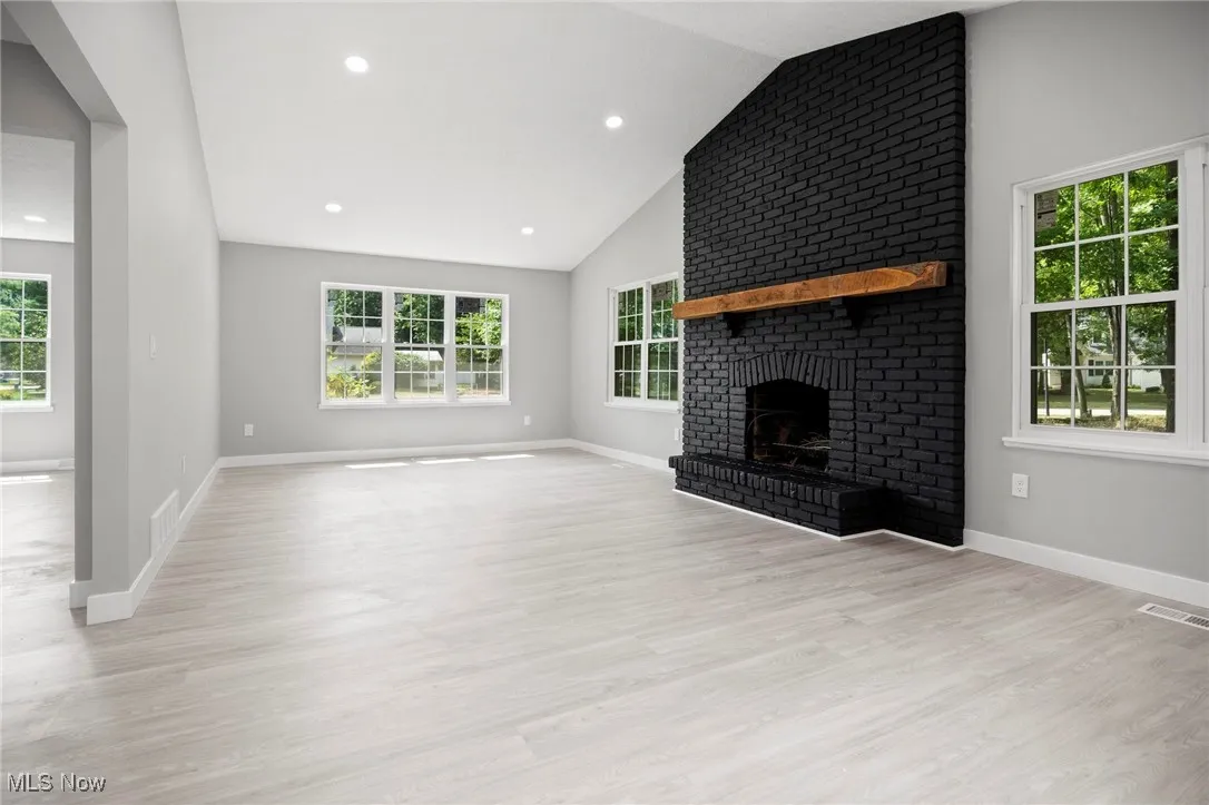 Unfurnished living room featuring healthy amount of natural light, a brick fireplace, light wood-style flooring, lofted ceiling, and recessed lighting