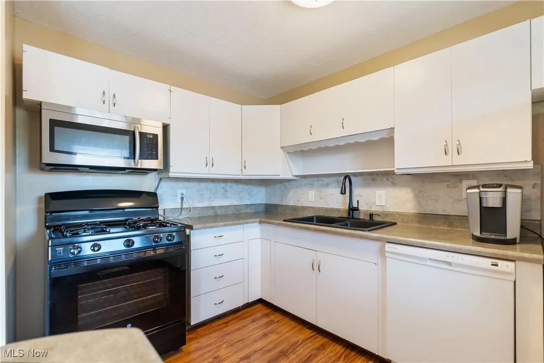 Kitchen with black gas range oven, dishwasher, stainless steel microwave, wood finished floors, and white cabinets