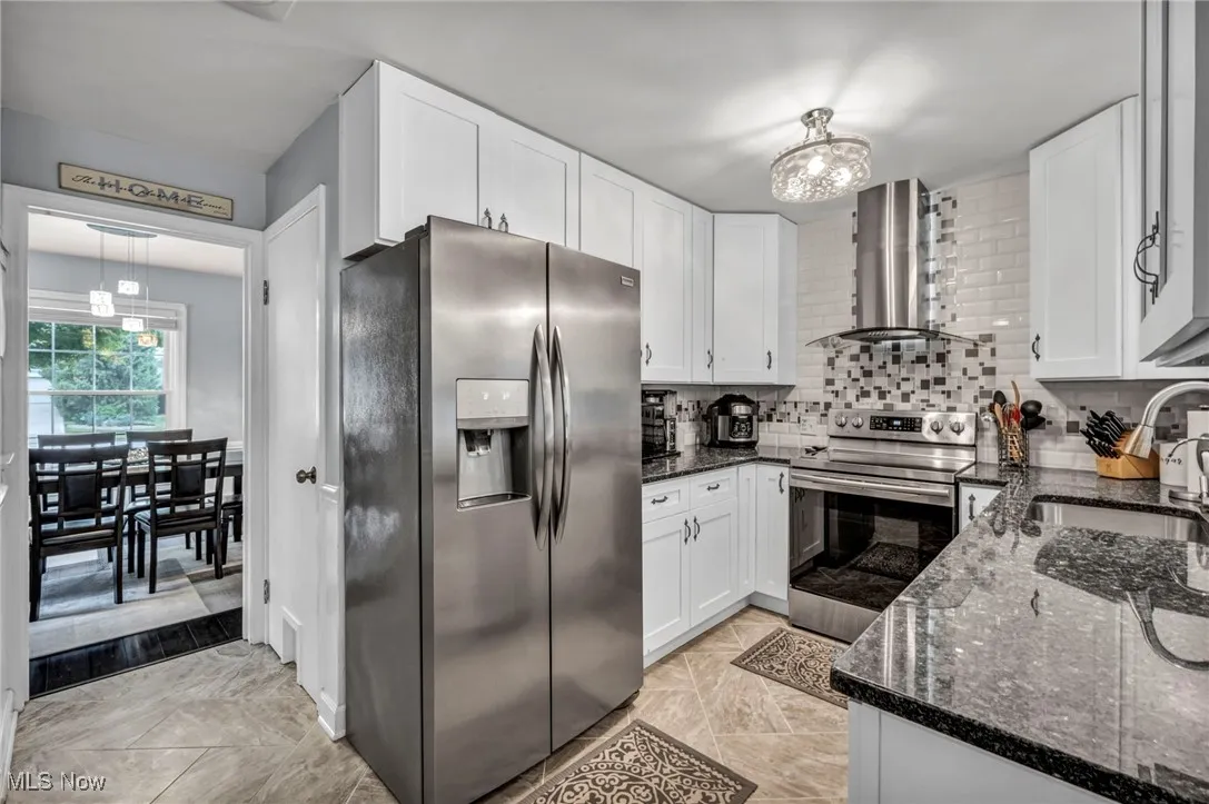 Kitchen featuring appliances with stainless steel finishes, wall chimney exhaust hood, white cabinetry, dark stone counters, and decorative backsplash