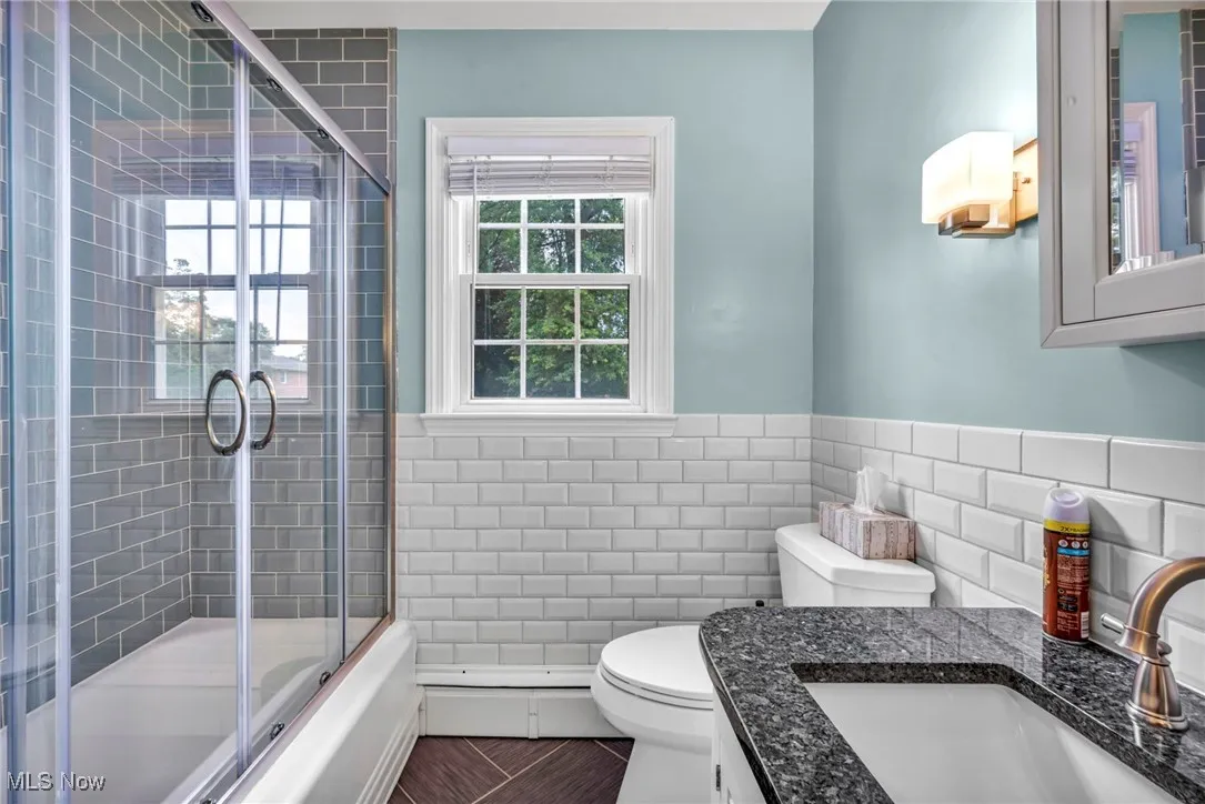 Bathroom with tile walls, vanity, combined bath / shower with glass door, wainscoting, and a baseboard heating unit