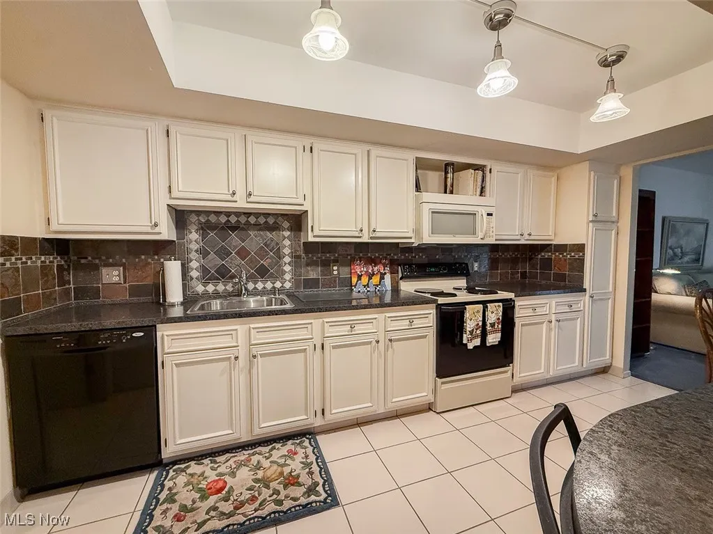 Kitchen featuring appliances, light cabinets with dark countertops, light tile patterned floors, and backsplash