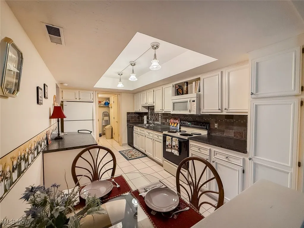 Kitchen featuring a raised ceiling, appliances, light cabinets with dark countertops, and tasteful backsplash