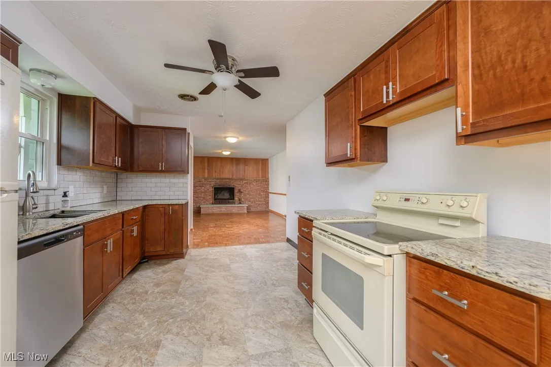 Kitchen featuring white range with electric cooktop, ceiling fan, stainless steel dishwasher, a brick fireplace, and light stone​​‌​​​​‌​​‌‌​‌‌​​​‌‌​‌​‌​‌​​​‌​​ counters