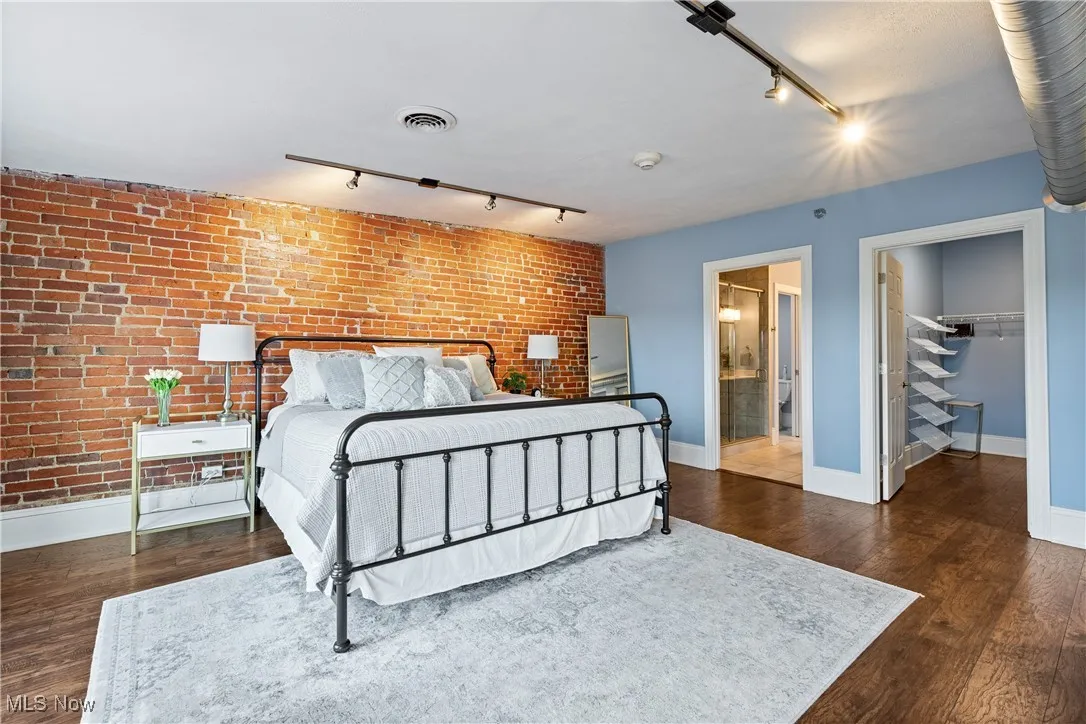 Bedroom with brick wall, track lighting, wood finished floors, a walk in closet, and ensuite bathroom