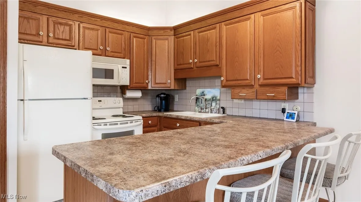 Kitchen with white appliances, a peninsula, tasteful backsplash, brown cabinetry, and light countertops
