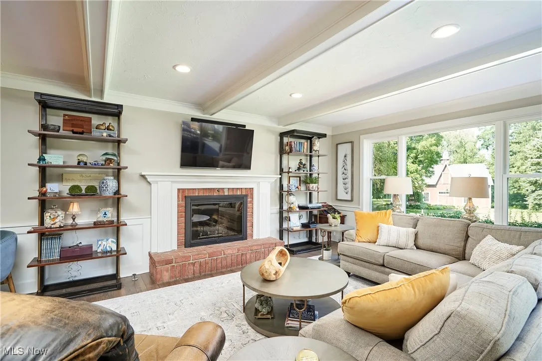 Living room featuring beam ceiling, wood finished floors, ornamental molding, a brick fireplace, and plenty of natural​​‌​​​​‌​​‌‌​‌‌​​​‌‌​‌​‌​‌​​​‌​​ light