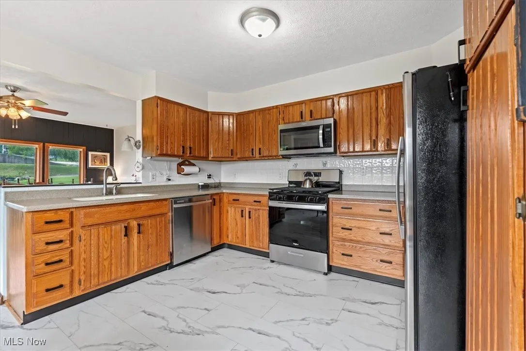 Kitchen featuring light marble finish floors, stainless steel appliances, brown cabinetry, and a ceiling fan. Kitchen is open to the living and dining room.