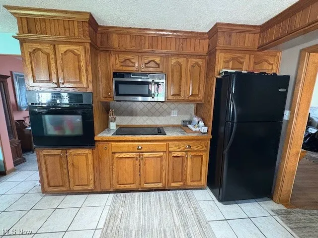 Kitchen featuring black appliances, brown cabinetry, light tile patterned floors, backsplash, and a textured ceiling