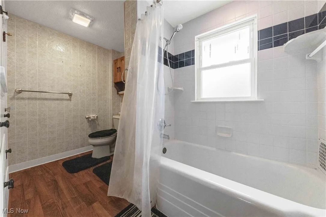 Bathroom featuring wood finished floors, shower / bathtub combination with curtain, and tile walls