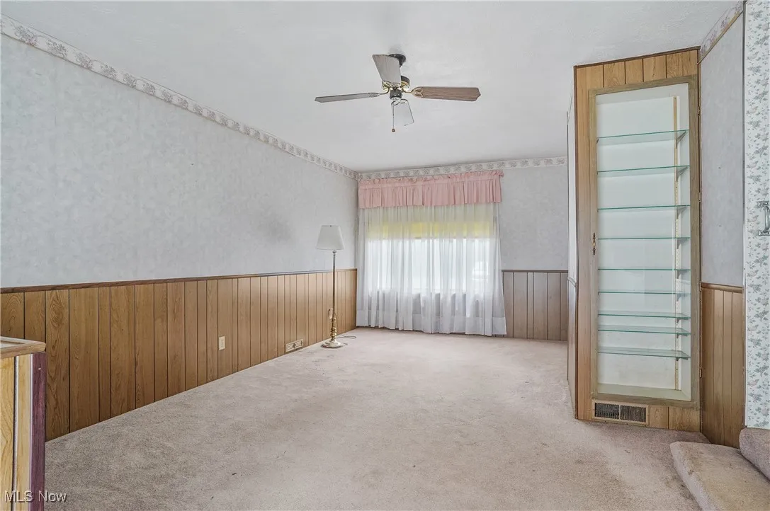 Carpeted empty room featuring wallpapered walls, wainscoting, a ceiling fan, and wood walls
