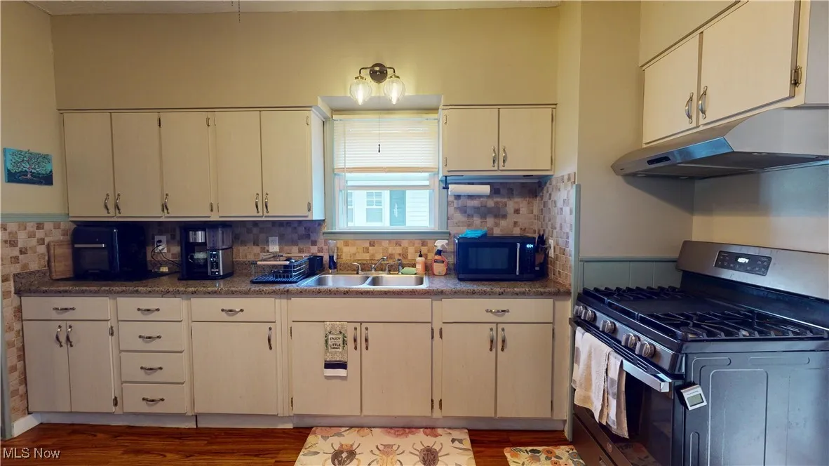 Kitchen featuring stainless steel gas range oven, under cabinet range hood, wood finished floors, black microwave, and backsplash