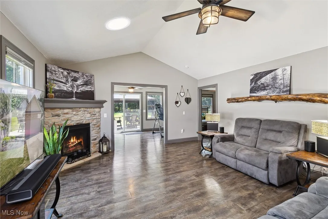 Living room featuring ceiling fan, plenty of natural light, wood finished floors, a stone fireplace, and vaulted ceiling