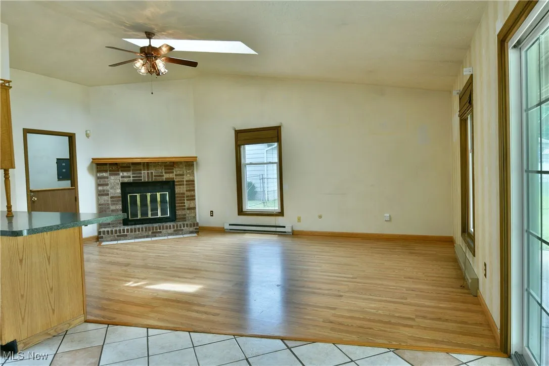 Unfurnished living room featuring vaulted ceiling, light wood-type flooring, ceiling fan, baseboard heating, and a brick fireplace