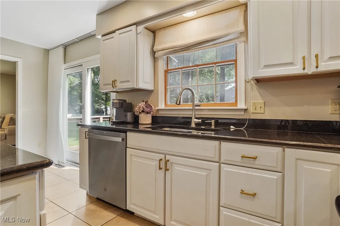 Kitchen with stainless steel dishwasher, light tile patterned flooring, white cabinets, and dark stone counters