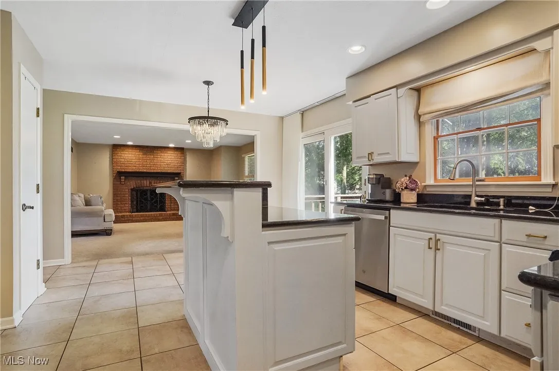 Kitchen with light tile patterned flooring, a fireplace, stainless steel dishwasher, hanging light fixtures, and white cabinetry