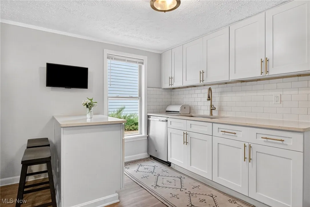 Kitchen featuring light wood finished floors, tasteful backsplash, light countertops, stainless steel dishwasher, and white cabinetry