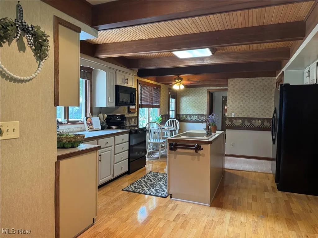 Large open Kitchen with wallpapered walls, wooden ceilings w/beams, black appliances, light wood finished floors, beam ceiling, and an island with sink