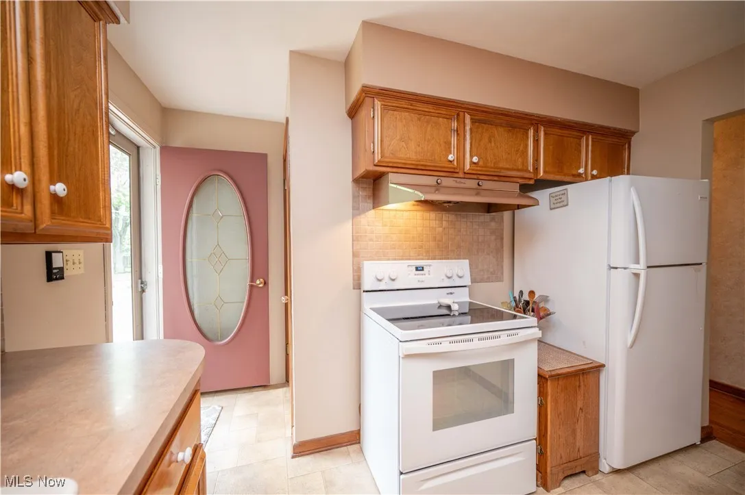 Kitchen featuring white appliances, brown cabinetry, under cabinet range hood, light countertops, and decorative backsplash