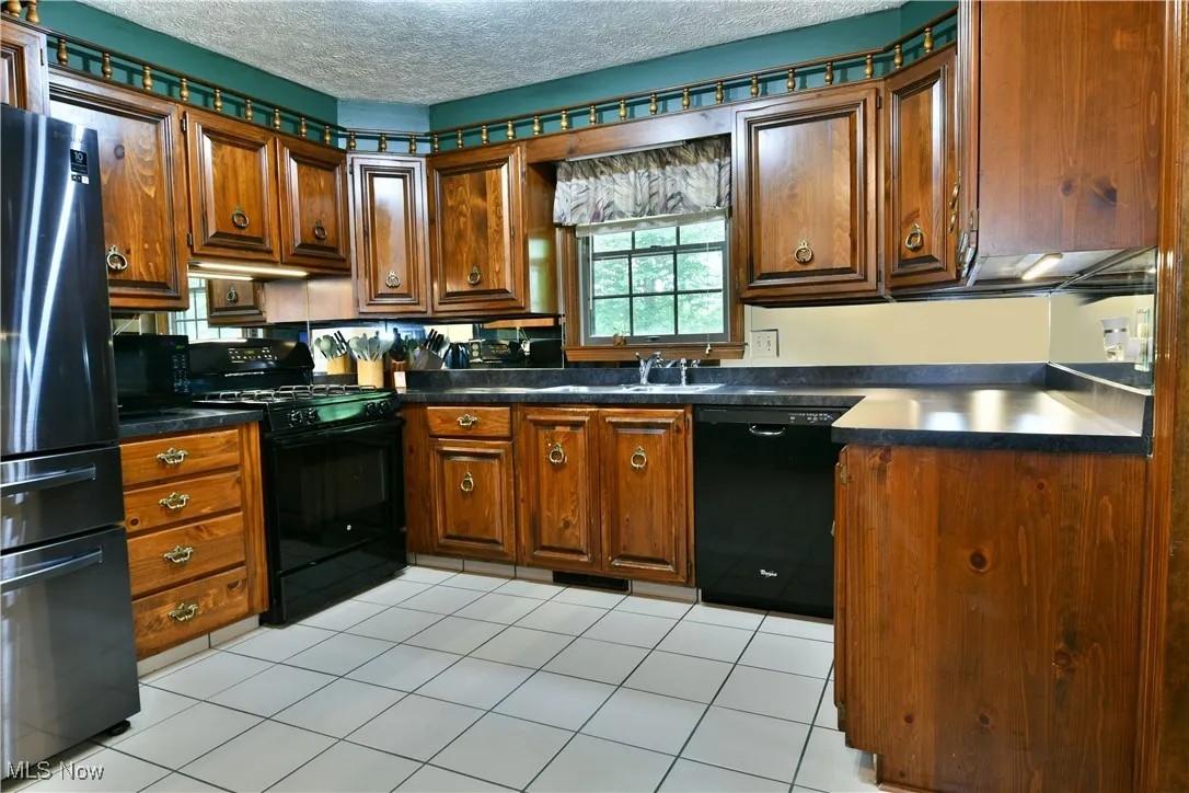 Kitchen with black appliances, a textured ceiling, dark countertops, light tile patterned flooring, and brown cabinets