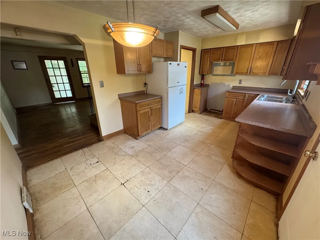 Kitchen featuring brown cabinetry, freestanding refrigerator, dark countertops, arched walkways, and open shelves