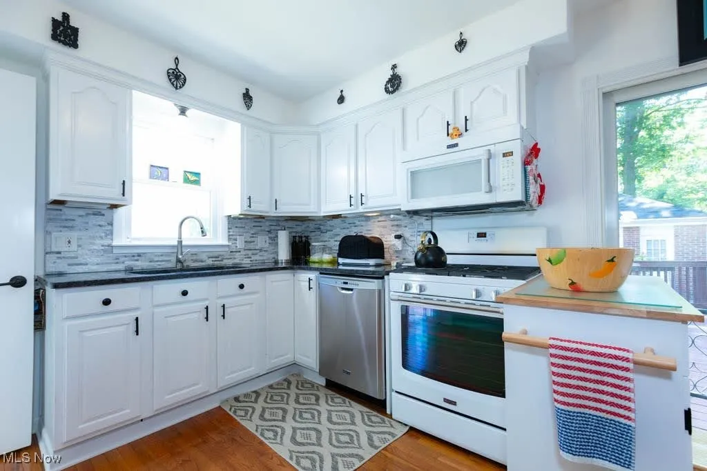 Kitchen featuring white appliances, white cabinets, decorative backsplash, light wood-type flooring, and dark countertops