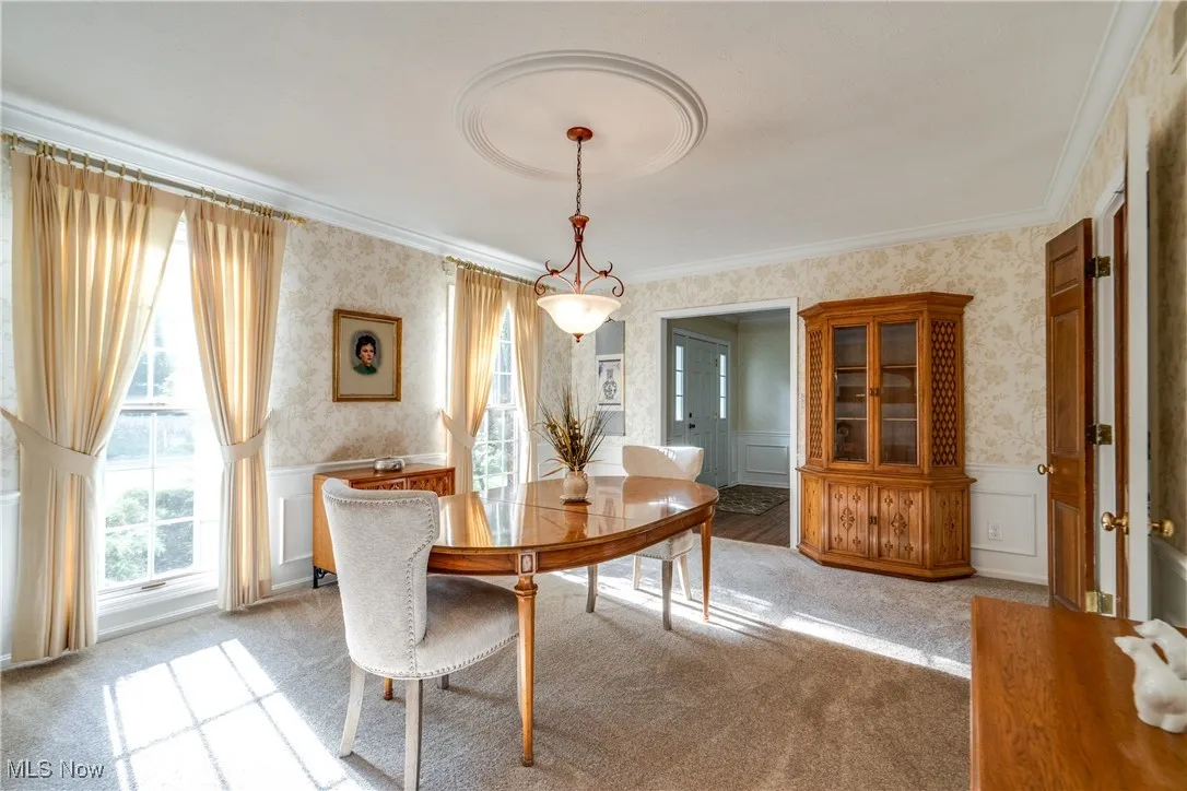 Dining space featuring wallpapered walls, light colored carpet, a wainscoted wall, crown molding, and a decorative wall