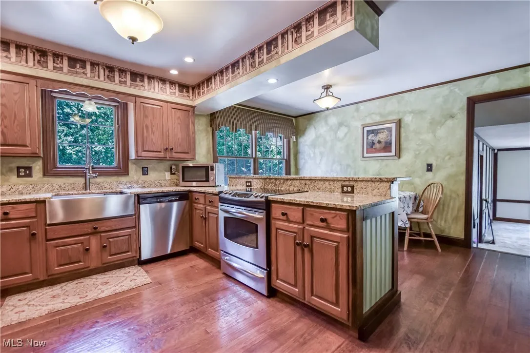 Kitchen with appliances with stainless steel finishes, dark wood-style flooring, a peninsula, light stone countertops, and brown cabinets
