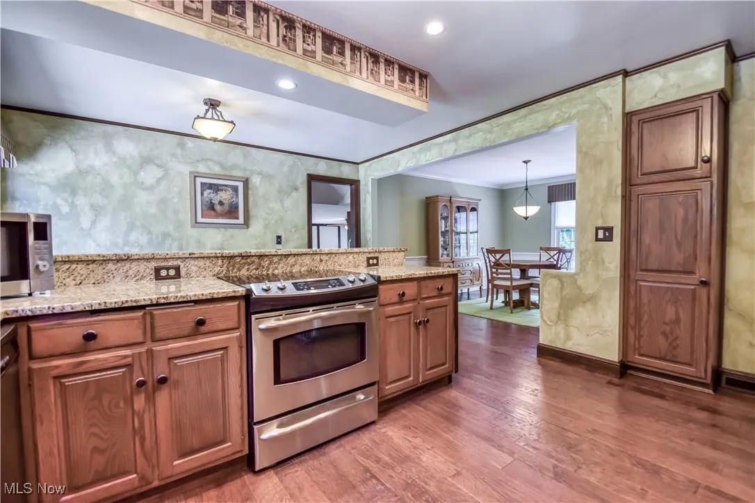 Kitchen with stainless steel appliances, crown molding, dark wood-type flooring, light stone countertops, and brown cabinetry