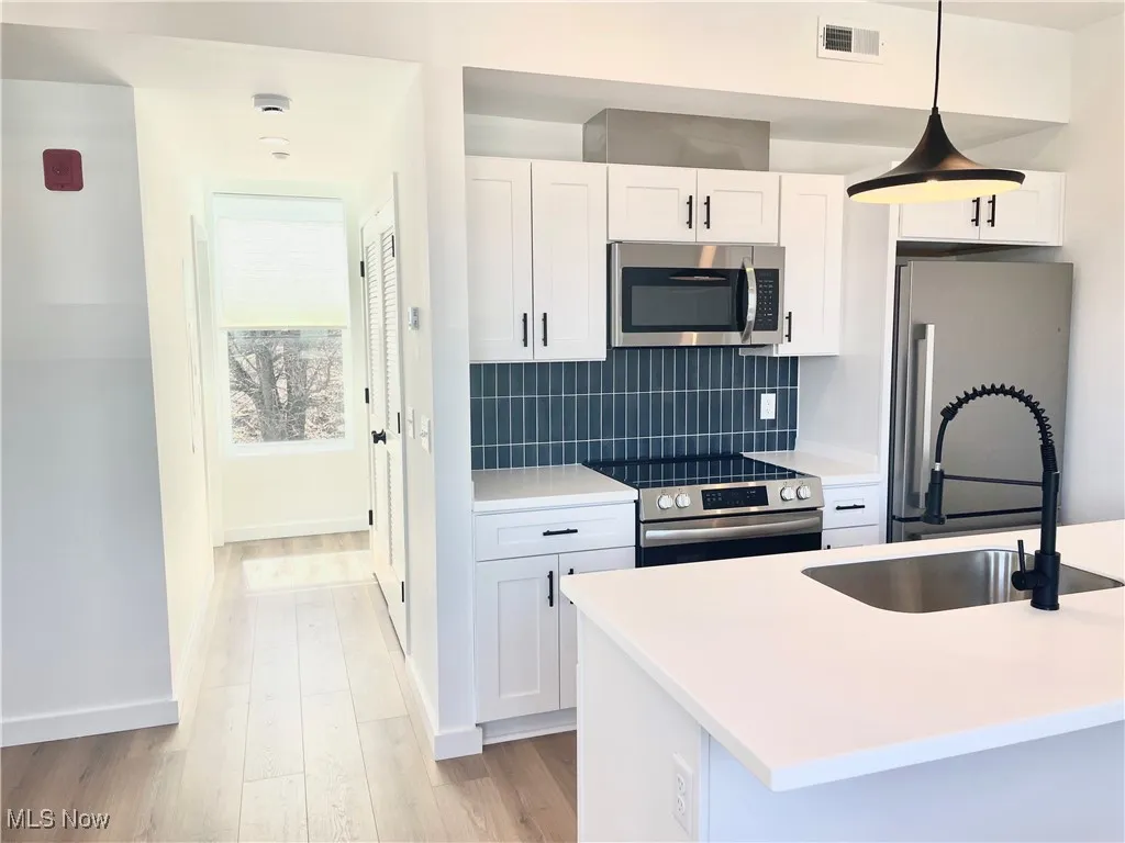 Kitchen featuring stainless steel appliances, white cabinets, decorative backsplash, light wood-style floors, and decorative light fixtures