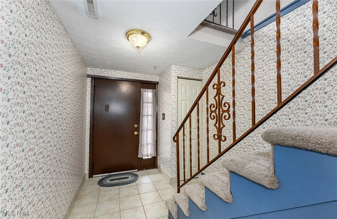 Tiled foyer entrance featuring wallpapered walls, stairs, and a textured ceiling