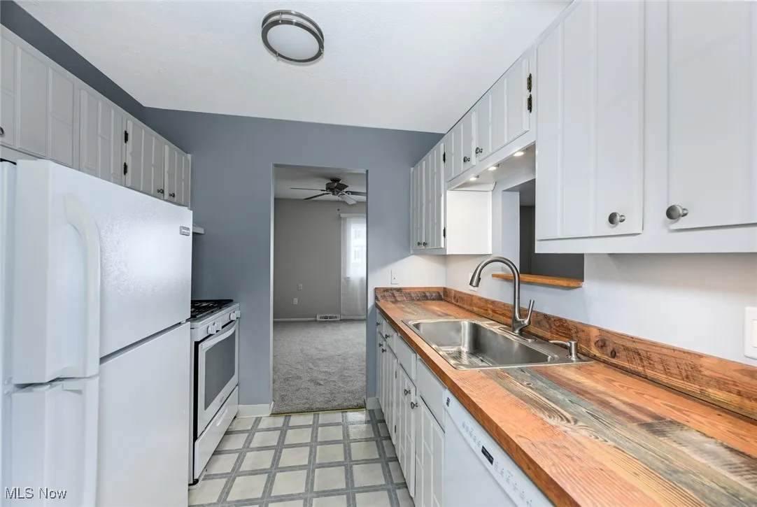 Kitchen featuring white appliances, a ceiling fan, light carpet, white cabinets, and wooden counters