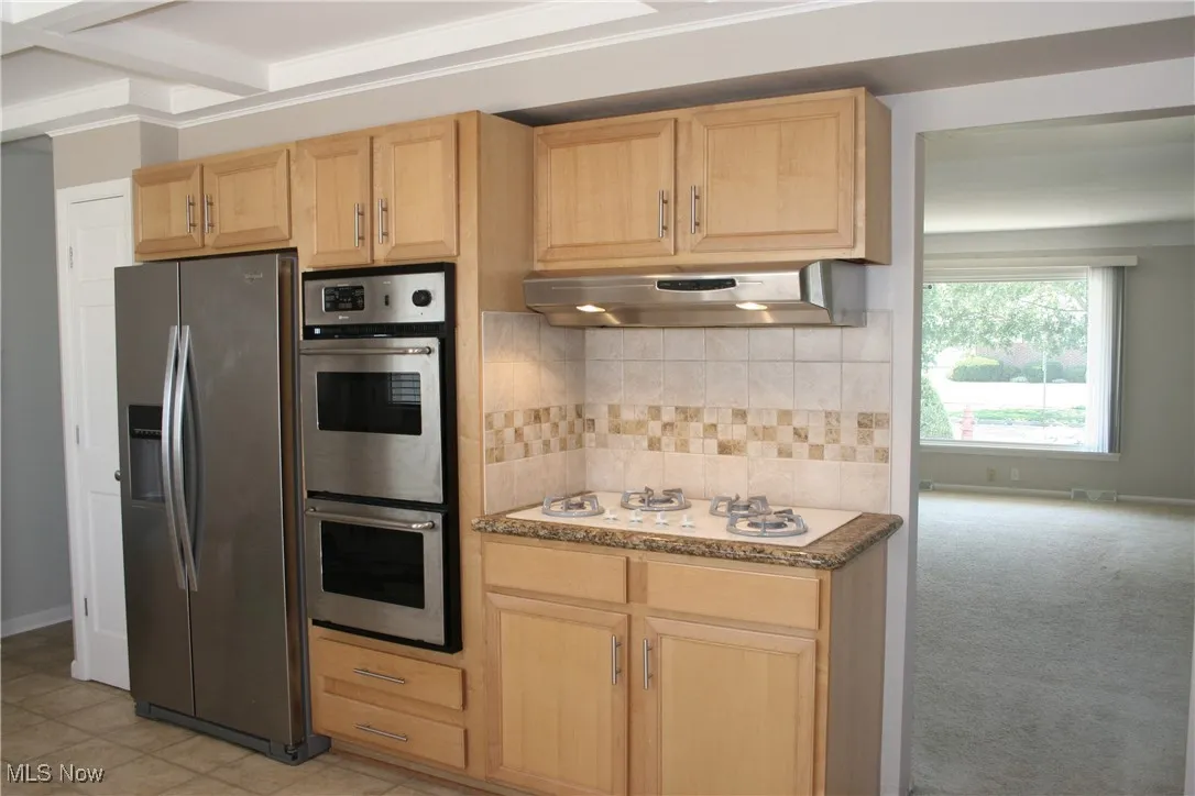 Kitchen featuring light brown cabinets, stainless steel appliances, under cabinet range hood, tasteful backsplash, and stone counters