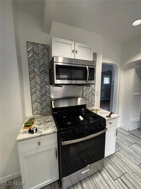 Kitchen with gas range oven, stainless steel microwave, arched walkways, white cabinets, and light stone counters