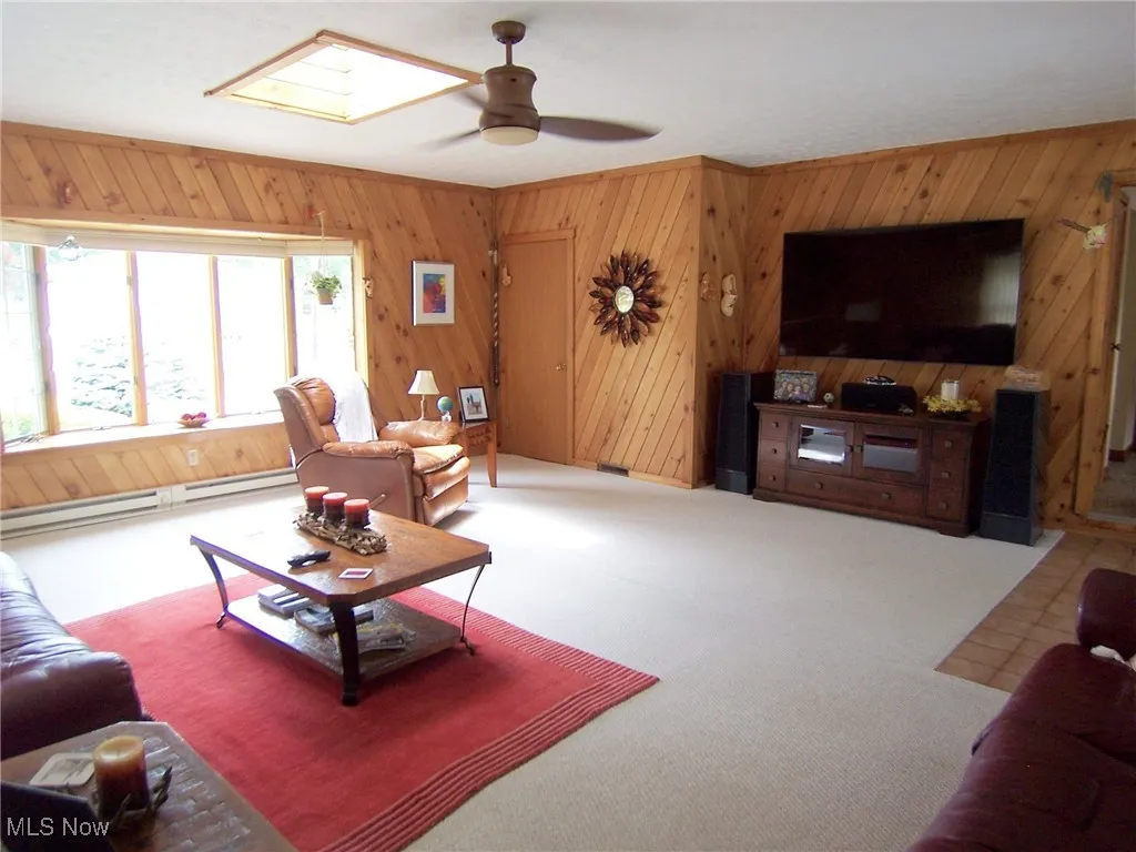 Living room with wood walls, a skylight, ceiling fan, a baseboard radiator, and carpet floors