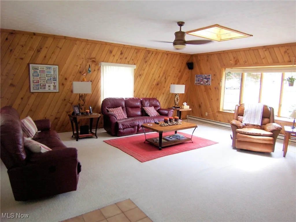 Carpeted living area with a ceiling fan, baseboard heating, a skylight, and wood walls