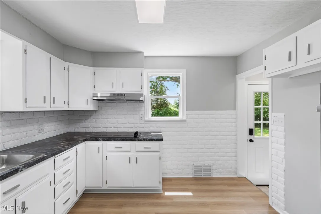 Kitchen featuring white cabinets, wood finished floors, dark countertops, under cabinet range hood, and tasteful backsplash