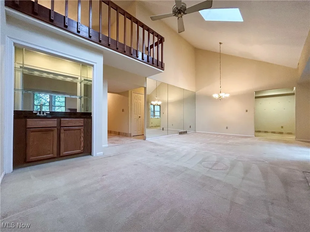 Living room featuring a wet bar, ceiling fan, a chandelier, view of dining area, a skylight, light carpet, and a high vaulted ceiling