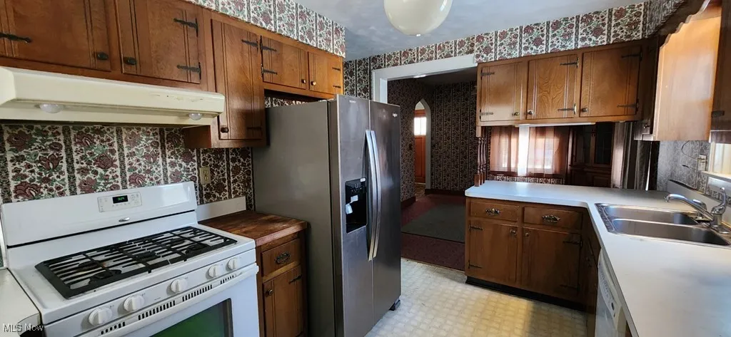Kitchen featuring wallpapered walls, white gas stove, under cabinet range hood, stainless steel fridge with ice dispenser, and light floors