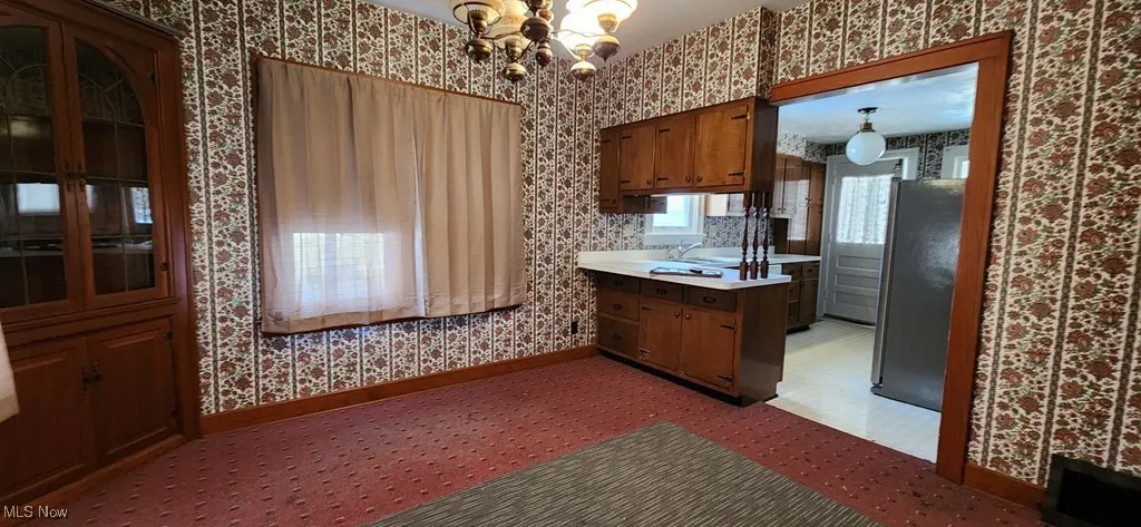 Kitchen with wallpapered walls, freestanding refrigerator, light countertops, a chandelier, and brown cabinetry