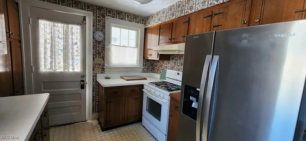 Kitchen featuring wallpapered walls, stainless steel fridge with ice dispenser, white range with gas stovetop, light flooring, and brown cabinetry