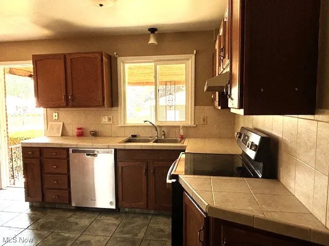 Kitchen featuring electric range oven, tile counters, stainless steel dishwasher, and backsplash